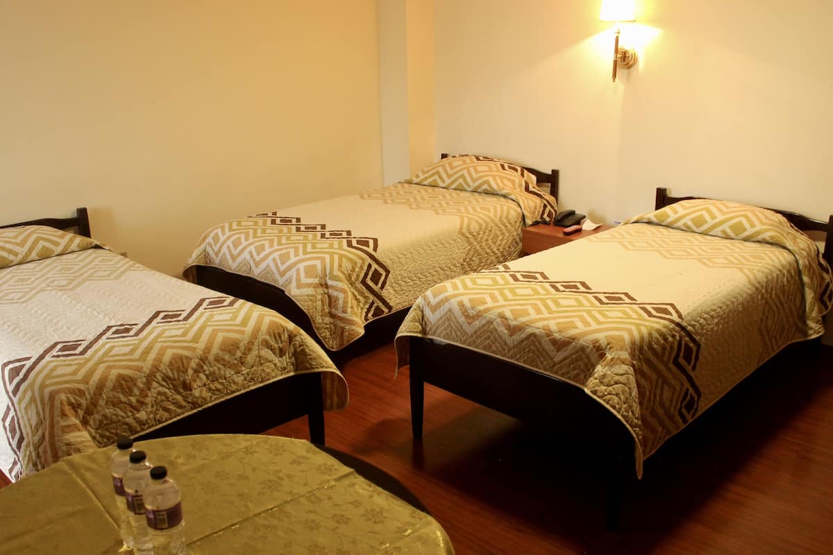 Une chambre d’hôtel avec quatre lits simples, chacun agrémenté de draps à motifs. On y trouve une petite table recouverte d’une nappe à fleurs, ainsi que des bouteilles d’eau. Une lampe fixée au mur assure l’éclairage.