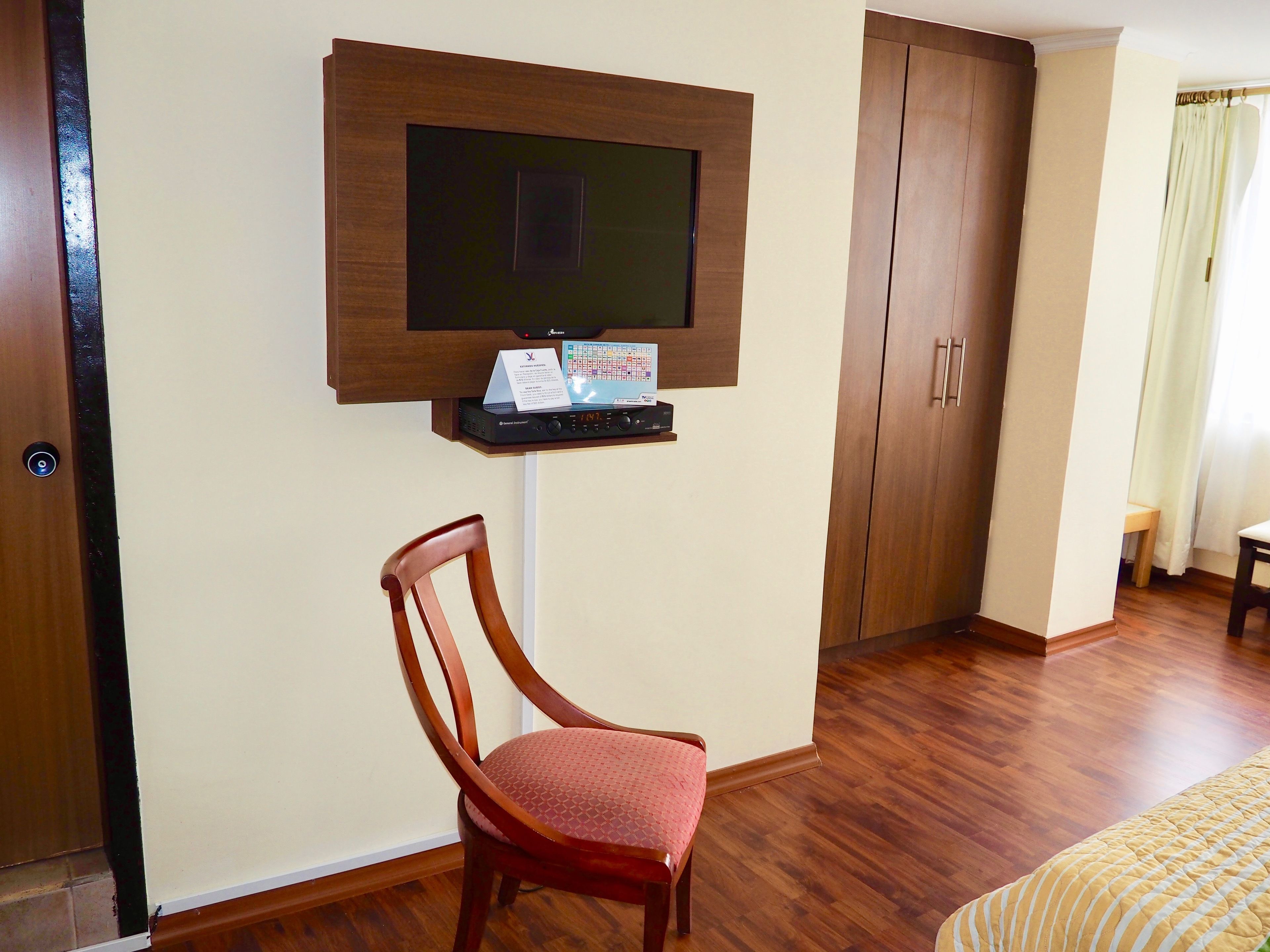 Quarto de hotel com piso de madeira, televisão fixada na parede, uma cadeira de madeira e cama com colcha amarela.