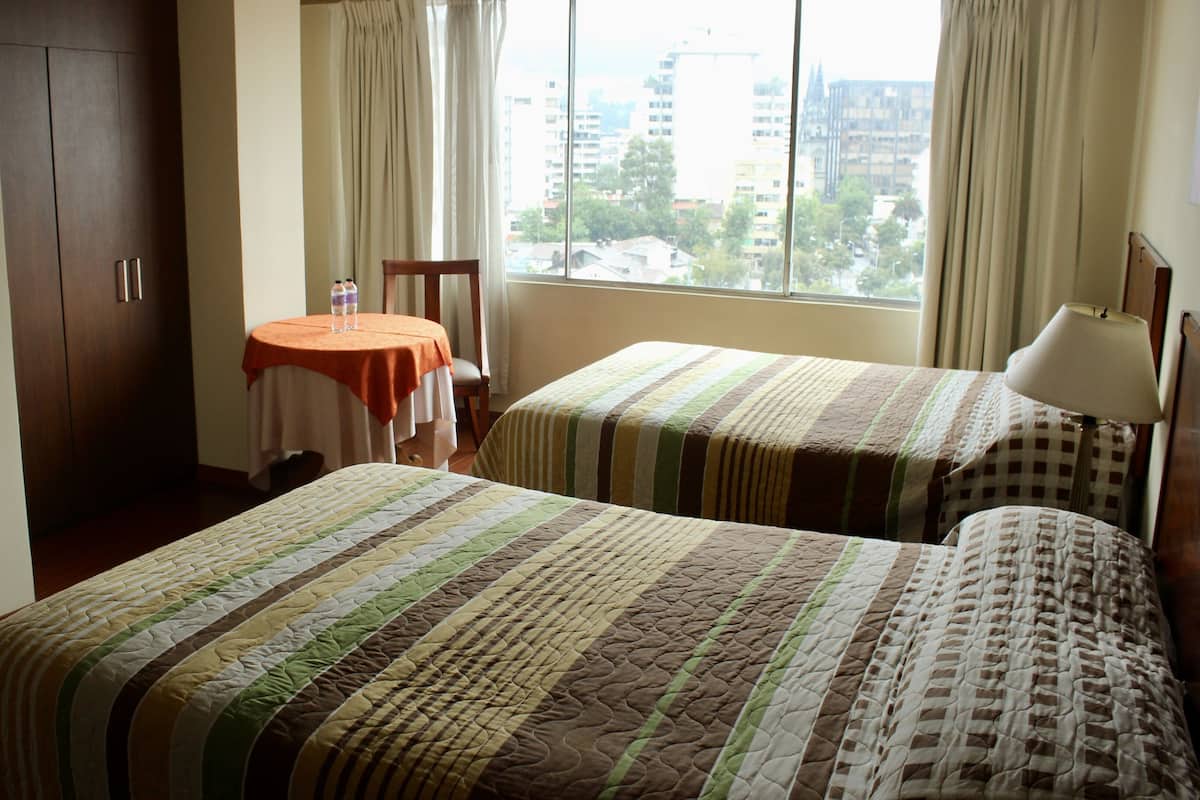 Une chambre d’hôtel avec deux lits, une petite table avec une chaise, une télévision et une fenêtre avec des rideaux.