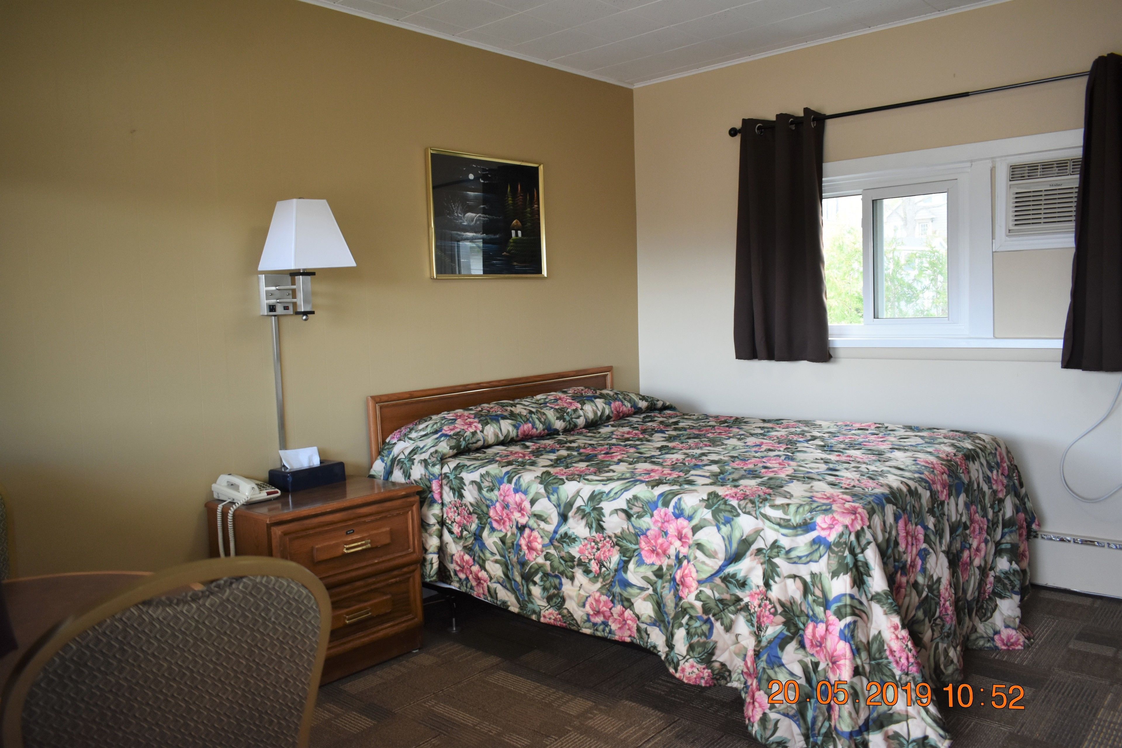 Habitación de hotel con cama, escritorio con lámpara, teléfono, silla, ventana con cortinas e imagen en un marco en la pared.
