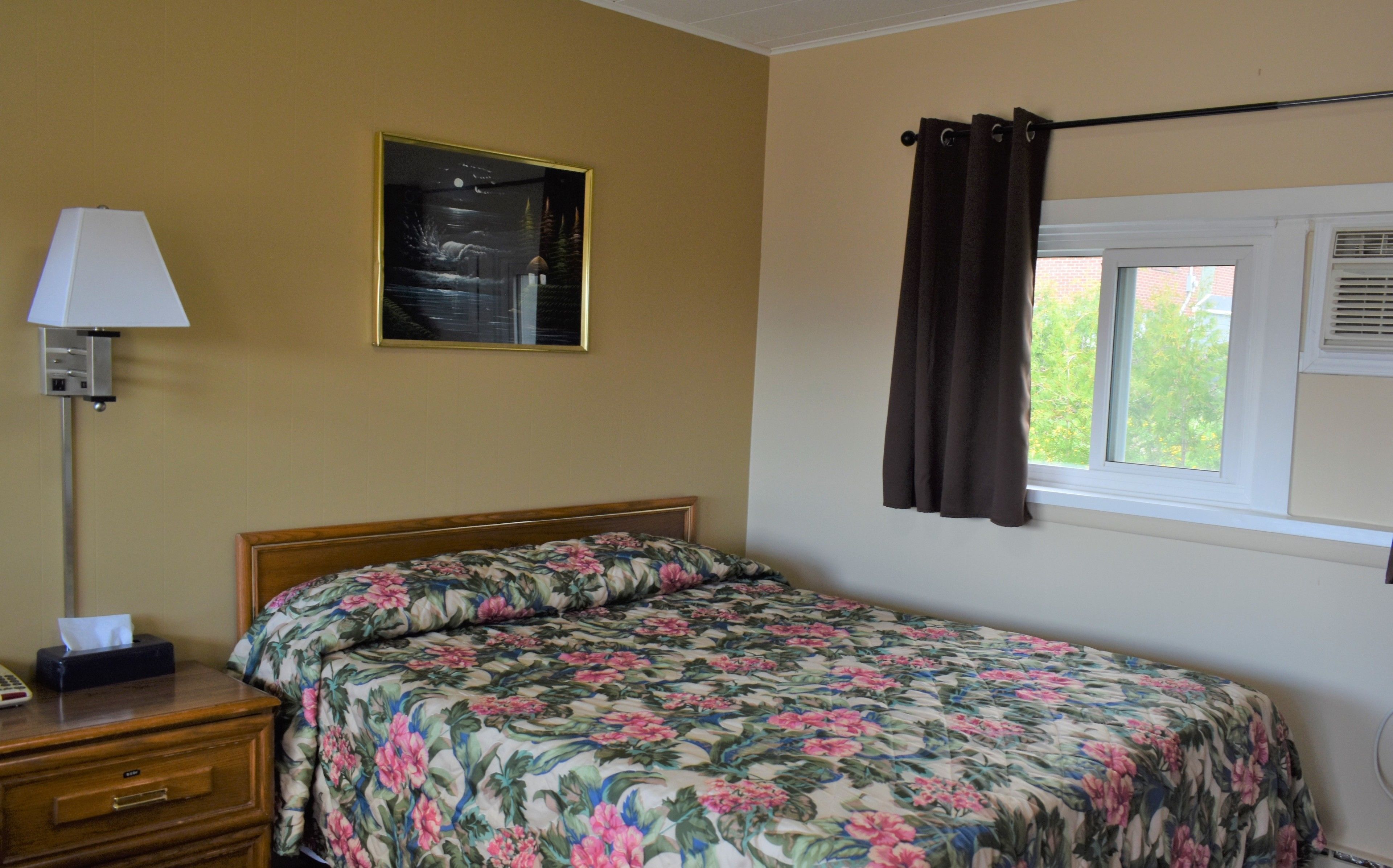 Una habitación de hotel con cama, mesita de noche, lámpara, un cuadro y ventana con cortinas.
