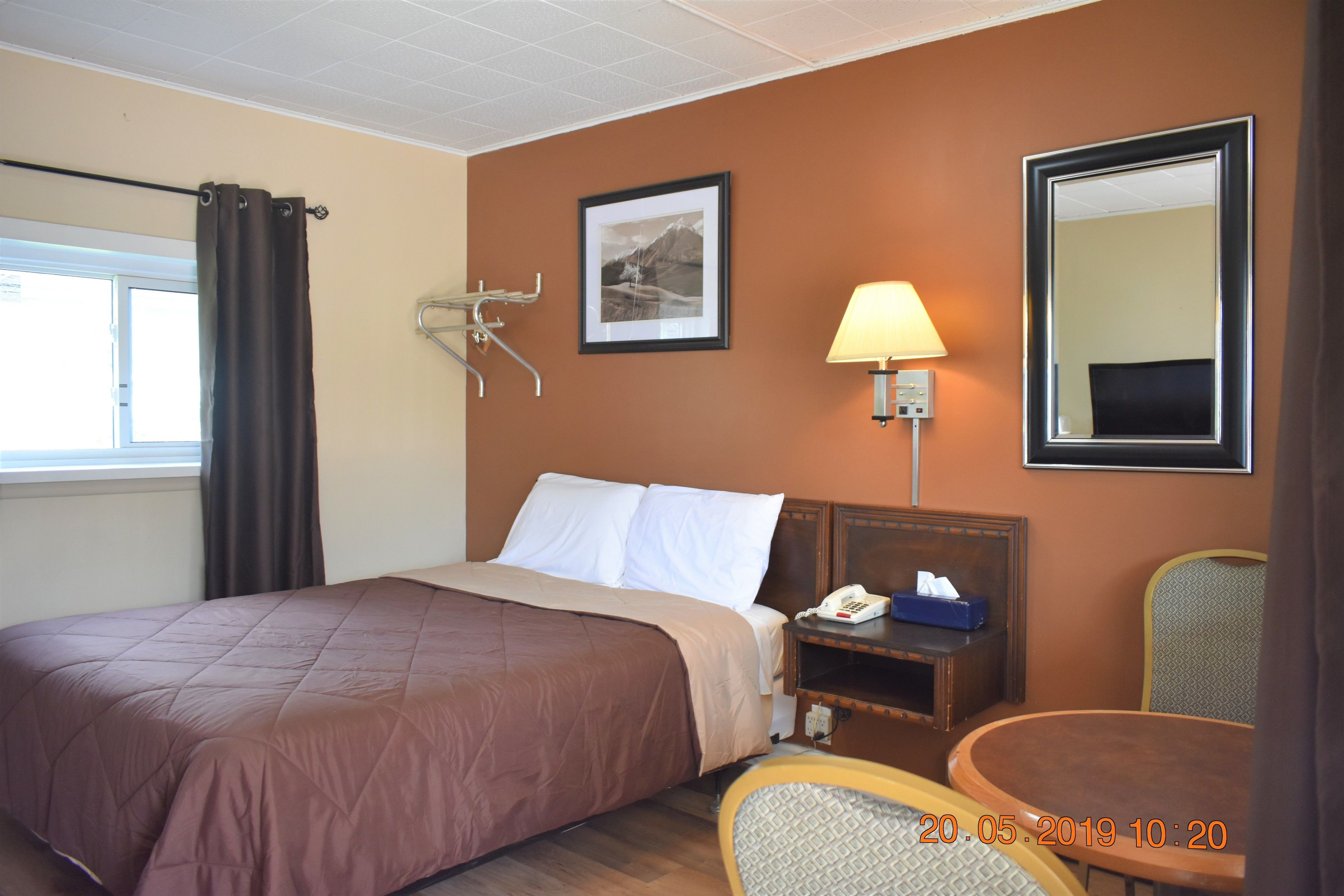 A hotel room with a bed, a bedside table with a lamp and phone, a chair, and a window with curtains.