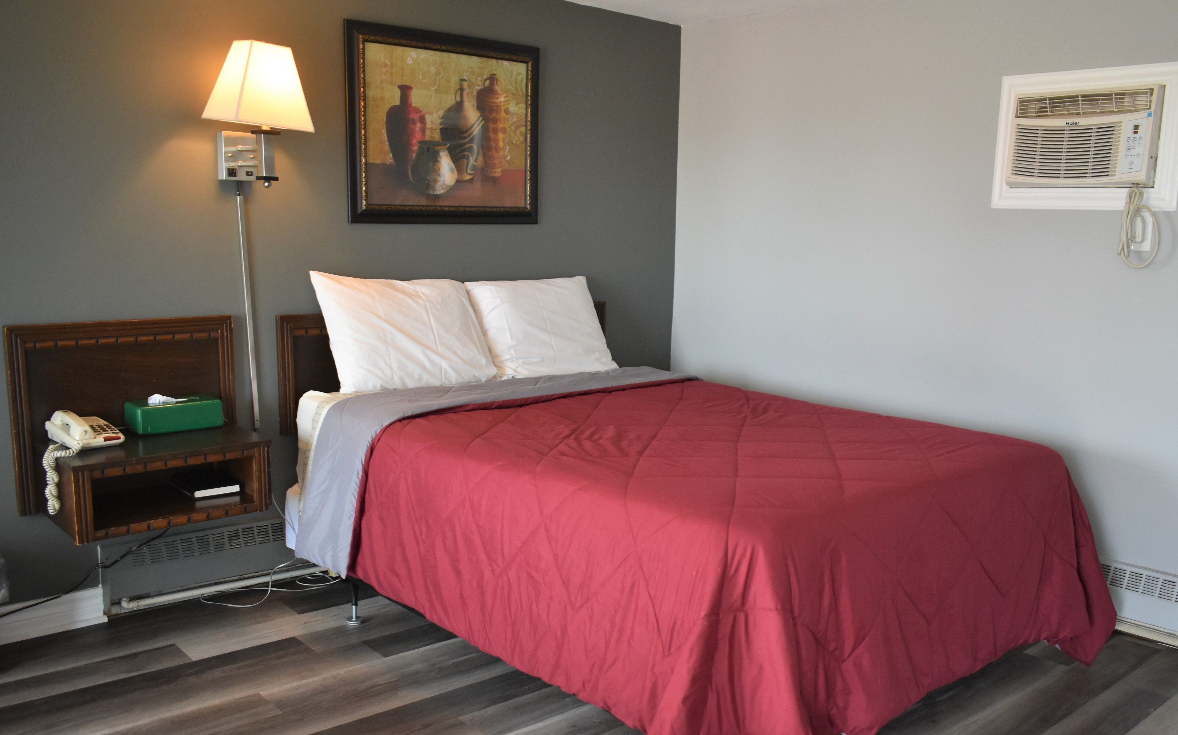 Habitación de hotel con cama, mesita de noche con lámpara, un cuadro en la pared y ventana con unidad de aire acondicionado.