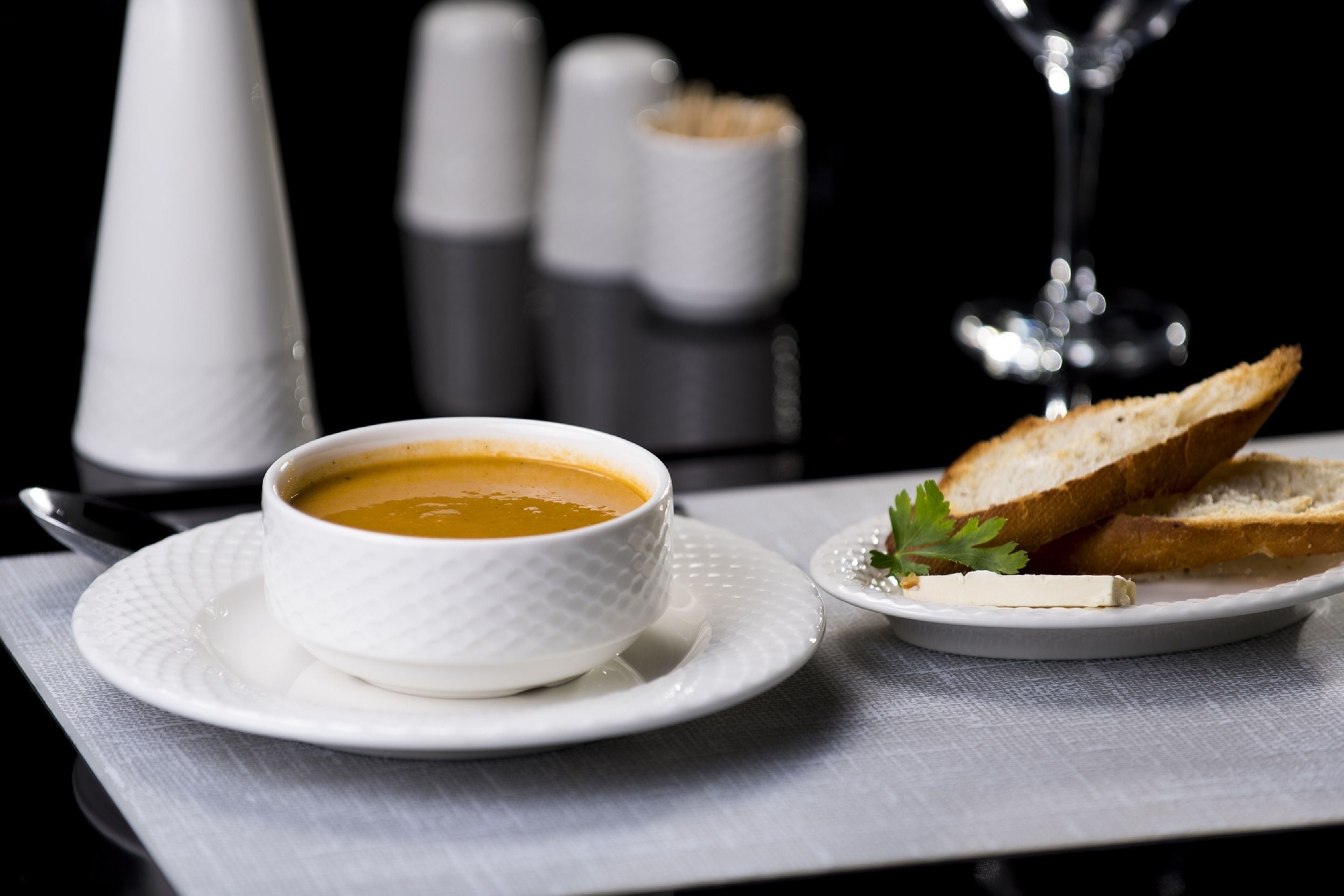 Eine weiße Schüssel mit Suppe auf einem weißen Teller, dazu ein Löffel, eine Scheibe Brot und eine kleine Servierschale mit Butter auf einer weißen Tischdecke.