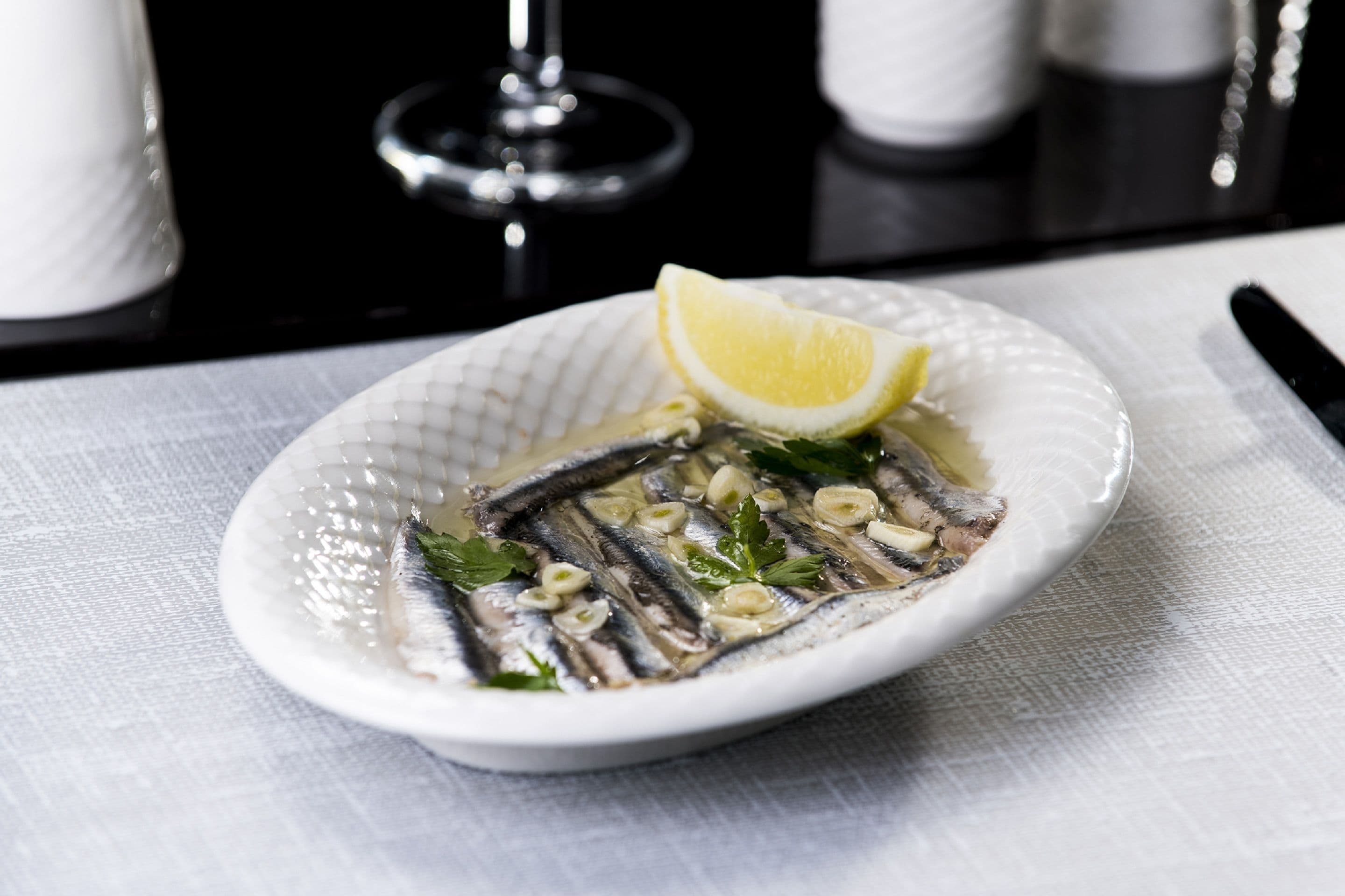 A white ceramic plate with a dish of sardines, garnished with lemon and parsley, on a white tablecloth.