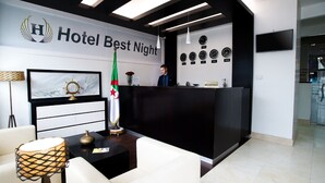 A hotel reception area with a reception desk, a receptionist, a wall clock, and a framed artwork. - Best Night (Algiers)