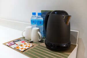 Habitación de hotel con cafetera, dos botellas de agua, una taza blanca, un hervidor blanco y una pequeña mesa con artículos de aseo.