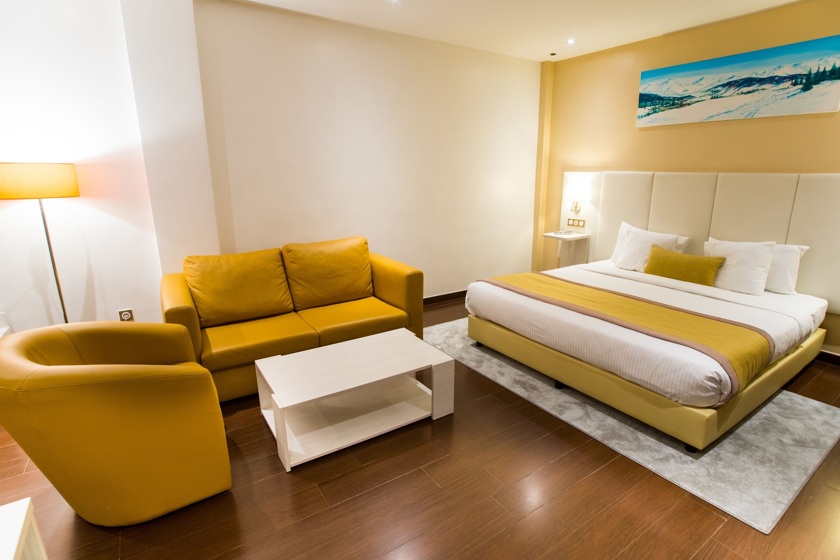 A modern hotel room with a bed, a yellow sofa, a white coffee table, and a painting on the wall.