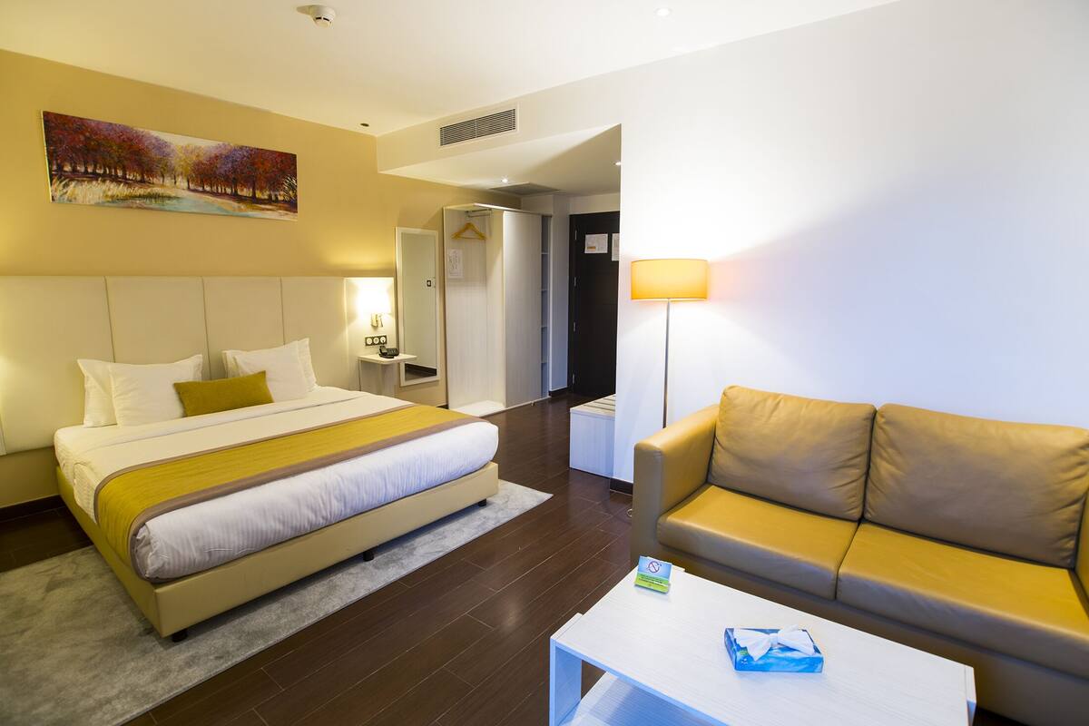 A hotel room with a bed, a sofa, a bedside table, a lamp, and a painting on the wall.