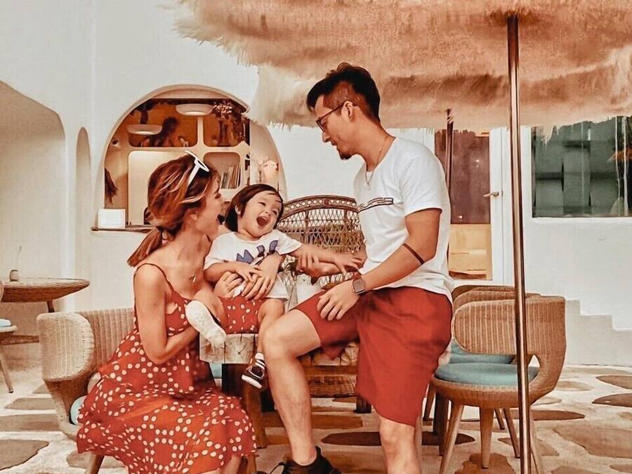 A family of three seated on a wicker chair under a large, fluffy umbrella in a room with a stone floor and white walls.