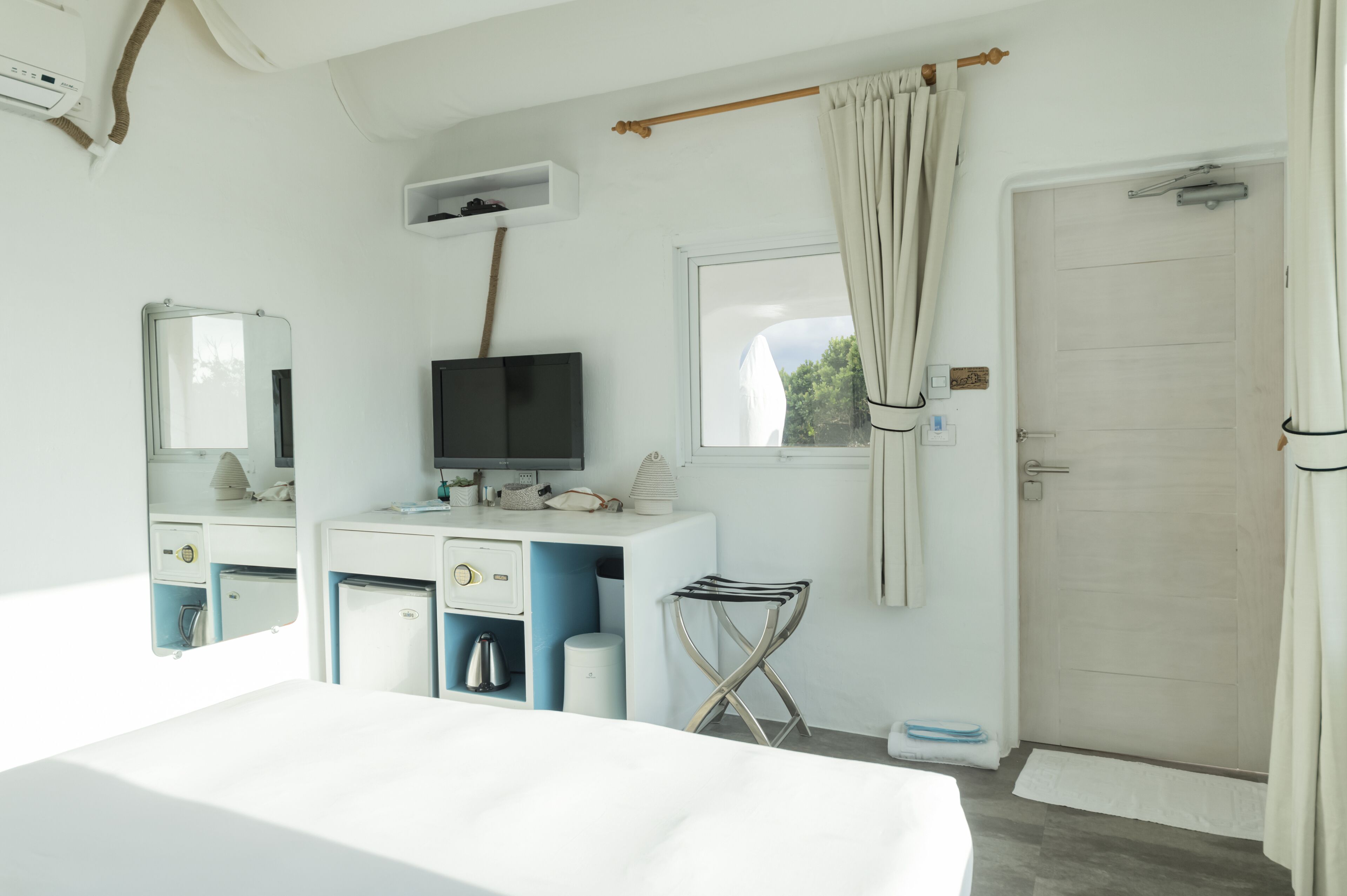 A hotel room with a bed, a TV, a desk with drawers, a small table, a window with curtains, and a door.