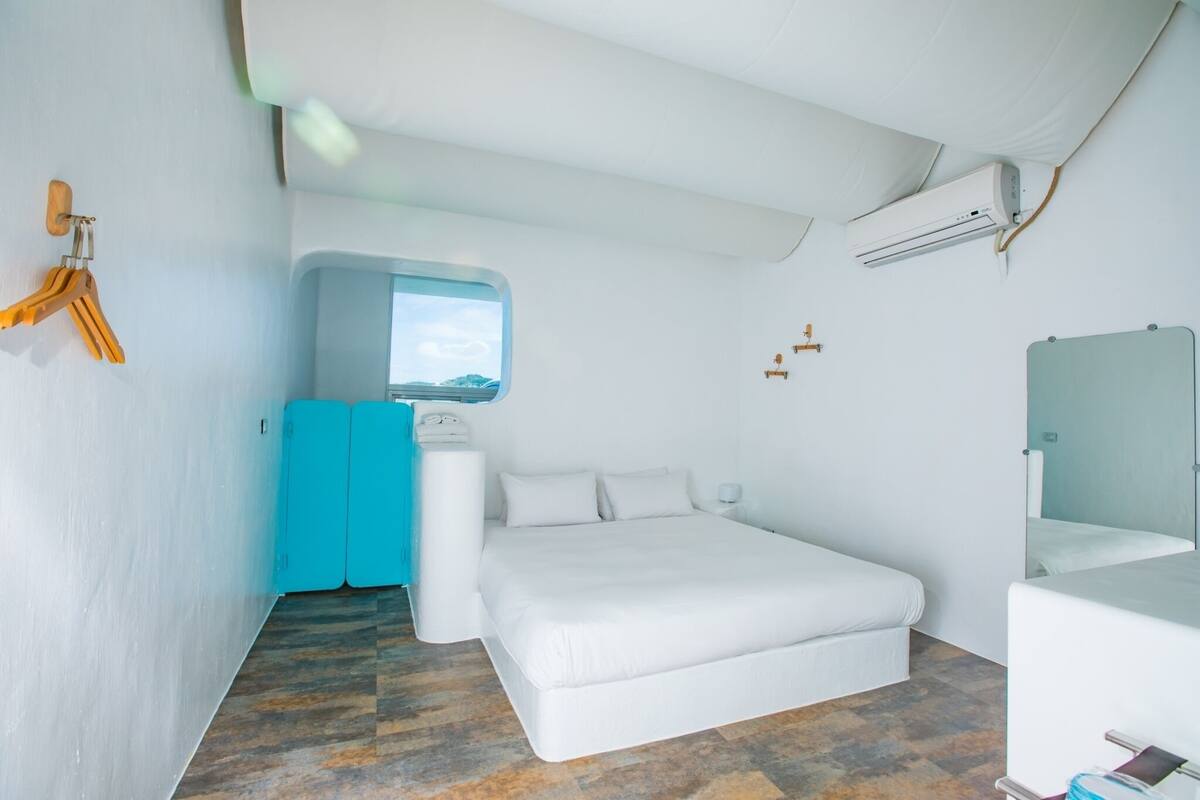 A modern hotel room with a bed, a small window, a blue cabinet, a white toilet, and an air conditioning unit.