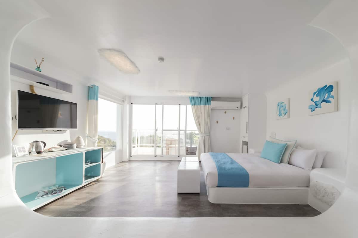 A modern hotel room with a large bed, a flat-screen TV, a minibar, and a balcony with a view.