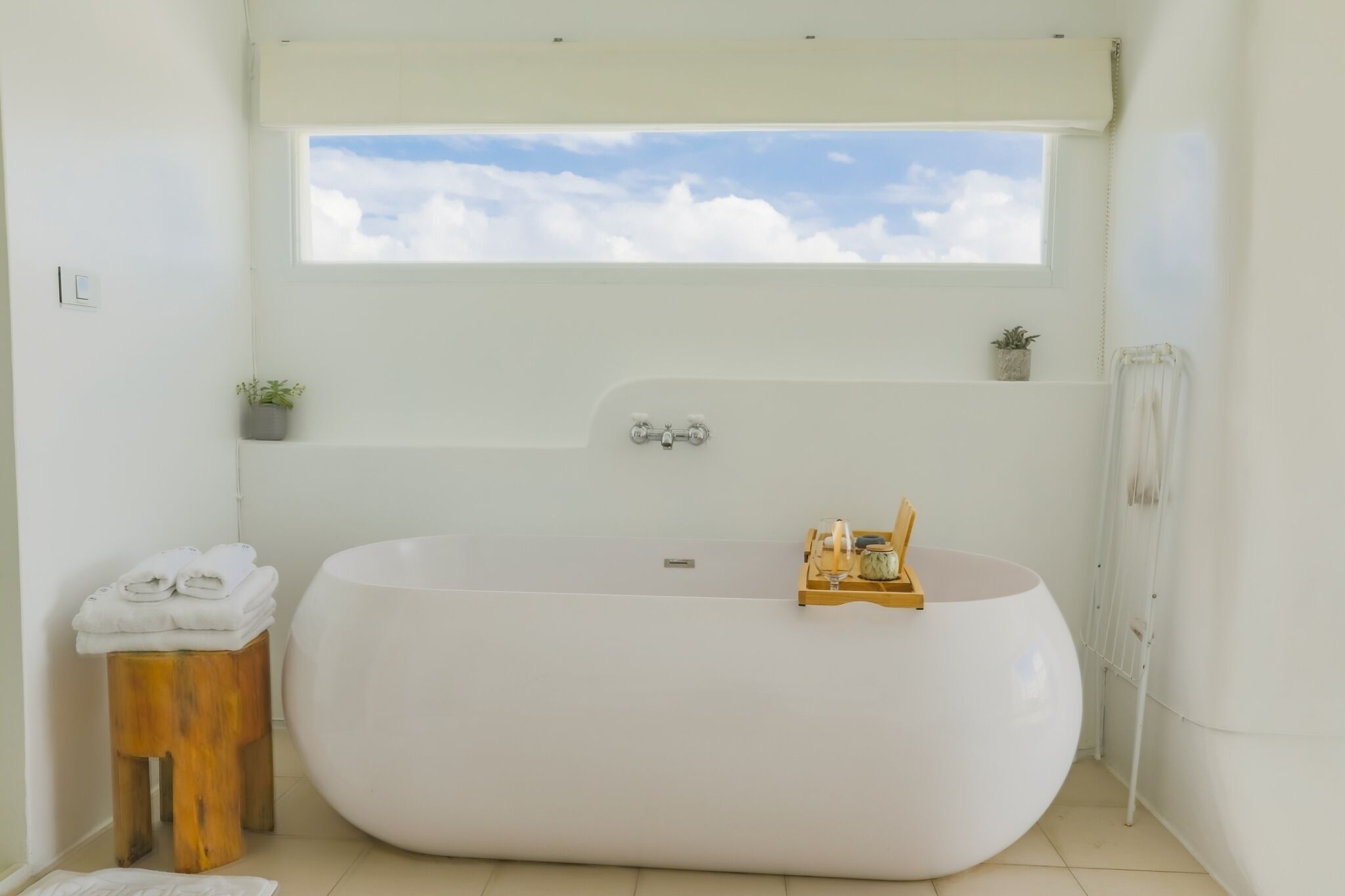 A modern bathroom with a large freestanding bathtub, a small wooden stool with towels, a window with a view of the sky, and a small shelf with a plant and a decorative item.
