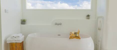 A modern bathroom with a large freestanding bathtub, a small wooden stool with towels, a window with a view of the sky, and a small shelf with a plant and a decorative item.