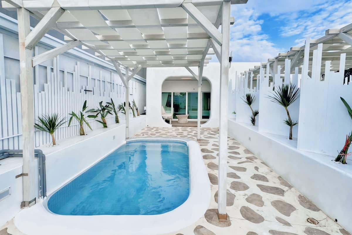 A swimming pool with a stone-tiled deck, surrounded by white walls and columns, and potted plants.