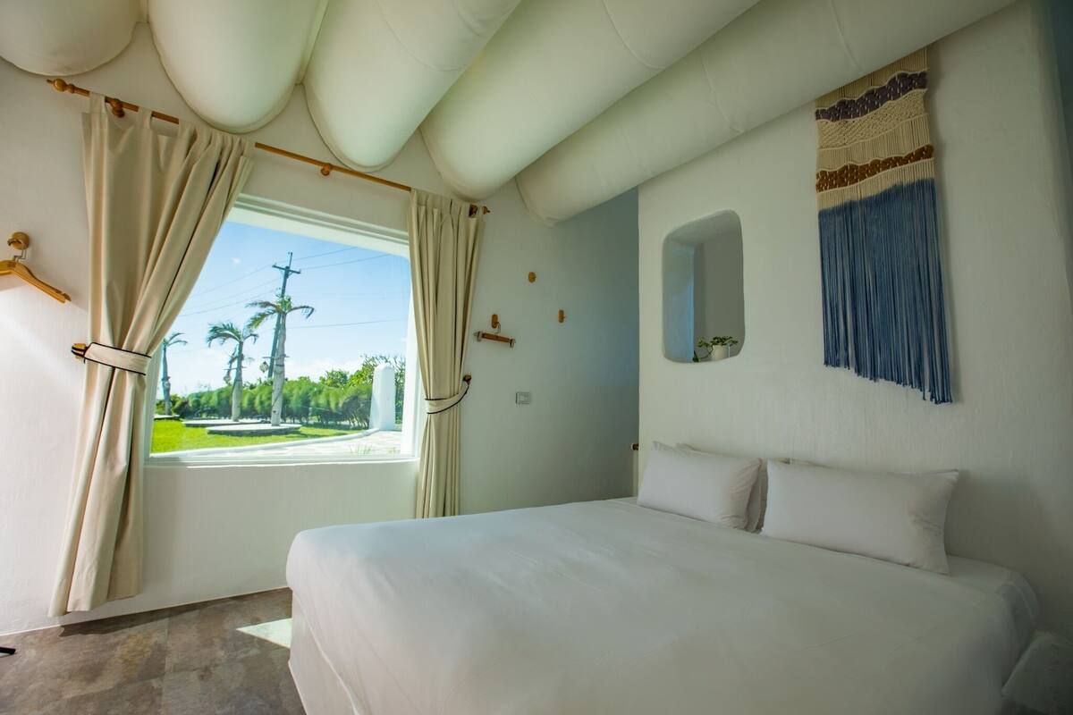 A hotel room with a bed, a window with curtains, a mirror, and a wall-mounted decorative piece.