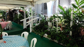 An outdoor patio area with a green carpet, white chairs, a table with a blue and white checkered tablecloth, and various potted plants.