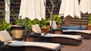 An outdoor seating area with wicker chairs and white cushions, surrounded by potted plants and umbrellas.