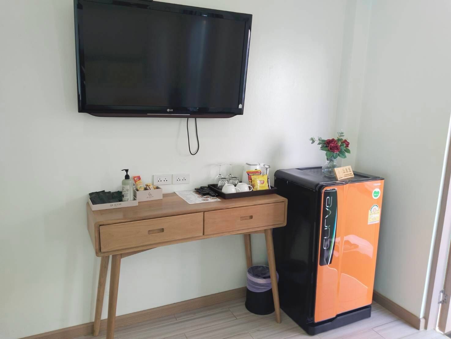 Una habitación con un televisor de pantalla plana en la pared, un escritorio de madera con cajones, un refrigerador naranja pequeño y un cubo de basura.