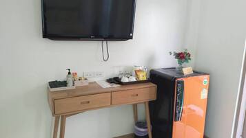 Una habitación con un televisor de pantalla plana en la pared, un escritorio de madera con cajones, un refrigerador naranja pequeño y un cubo de basura.