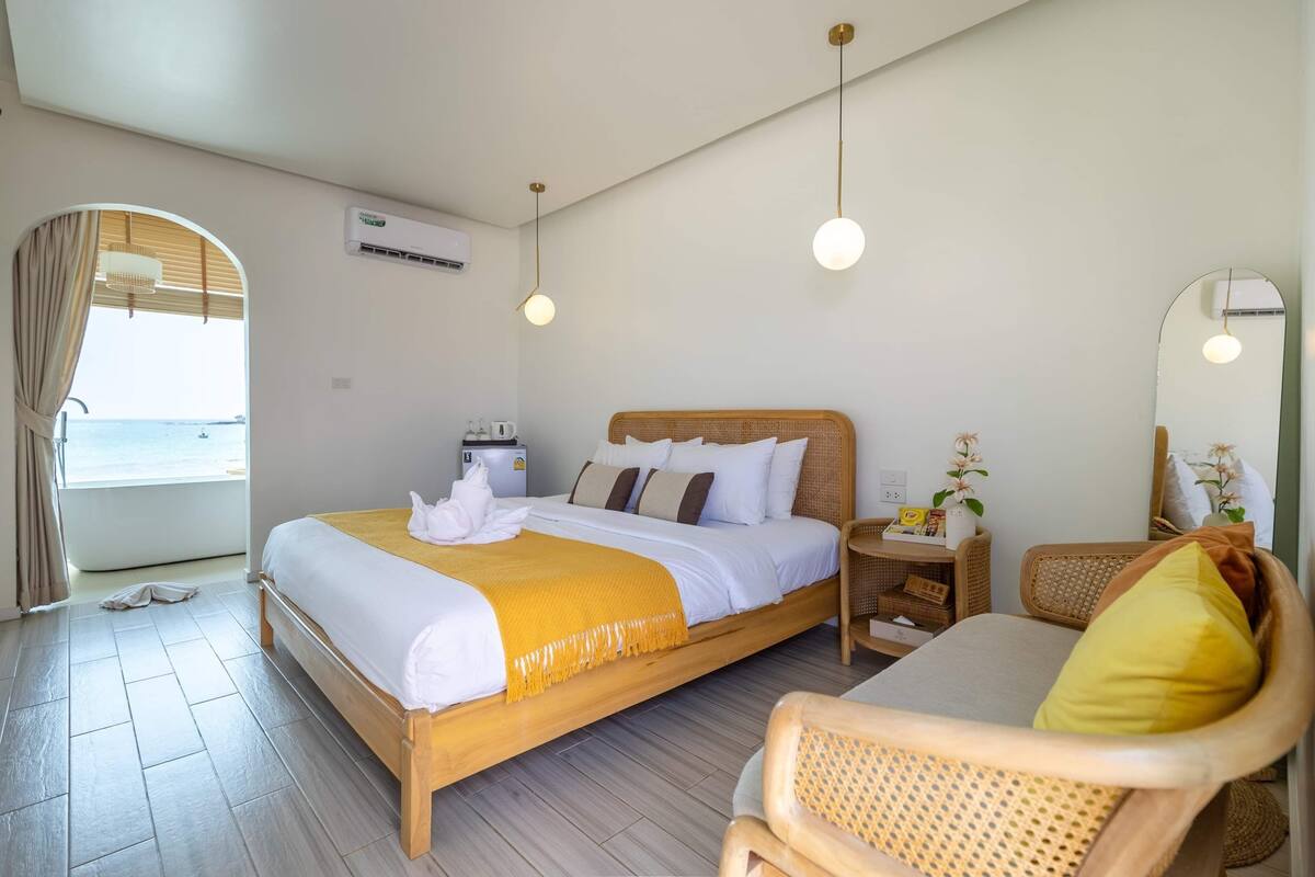 A hotel room with a bed, a chair, a bedside table, an air conditioner, and a view of the sea.