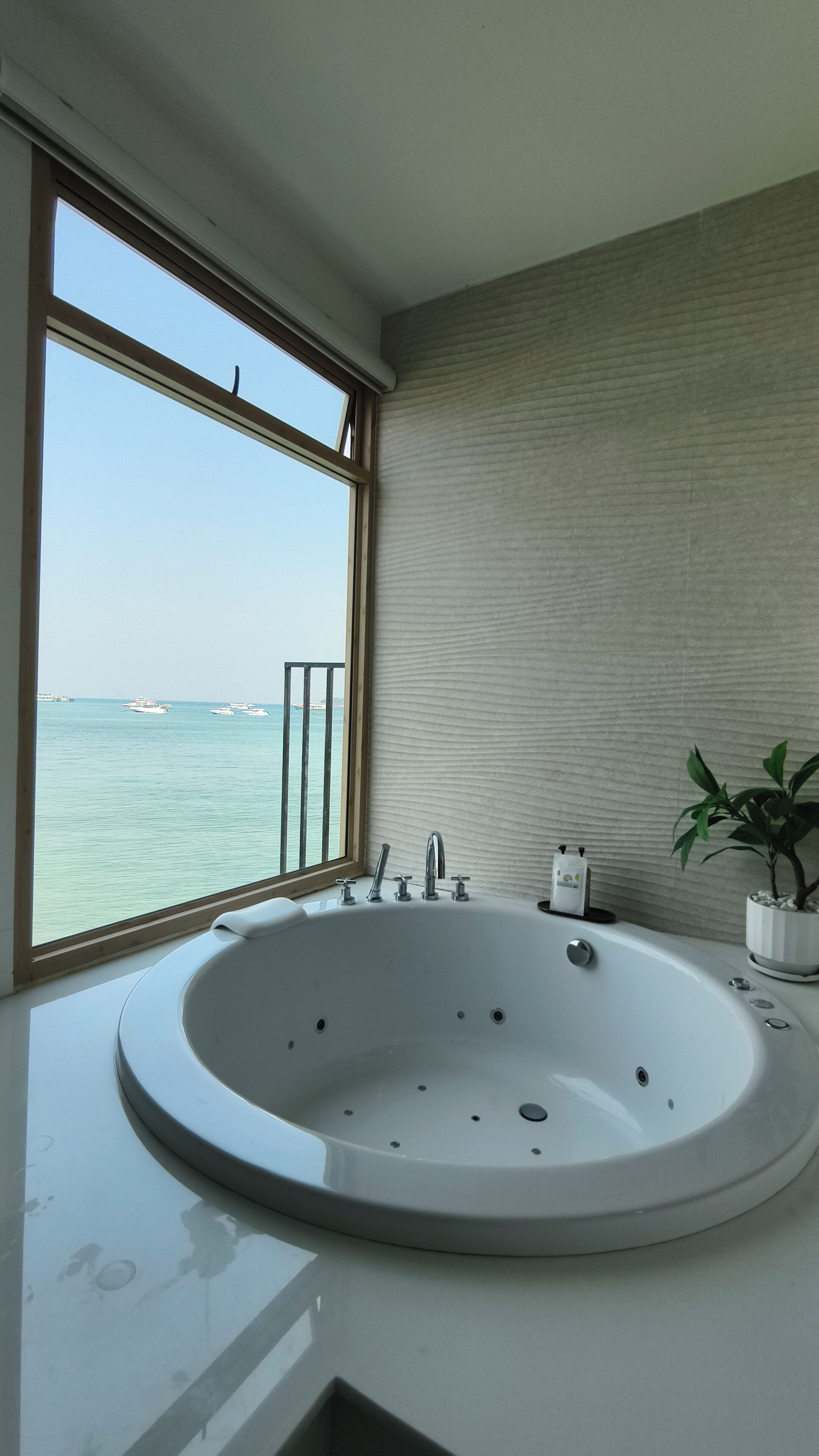 A modern bathroom with a large bathtub, a window with a view of the sea, and a potted plant.
