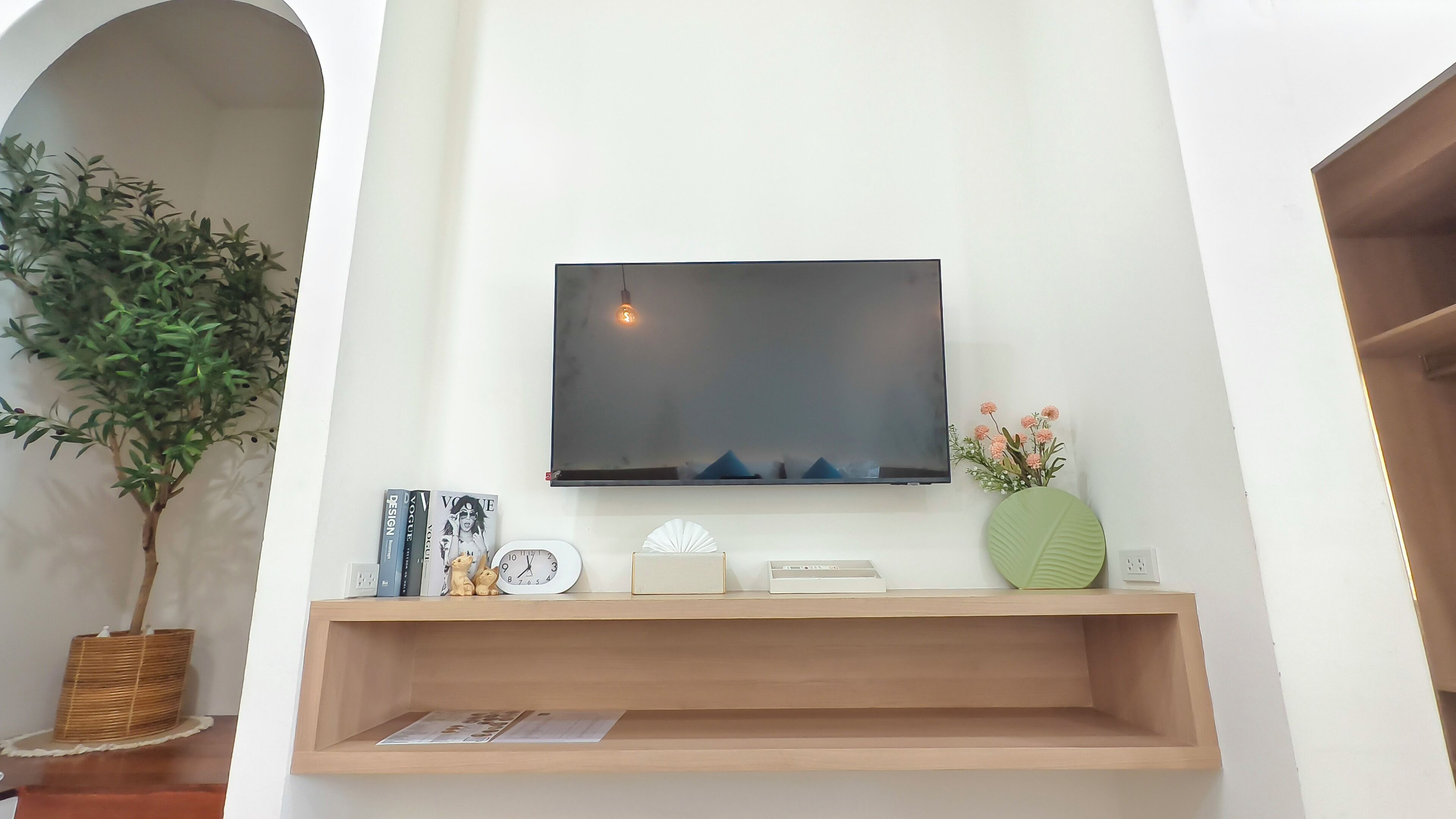Una sala de estar con un televisor montado, un estante de madera con objetos decorativos, una planta en maceta y un reloj de pared.