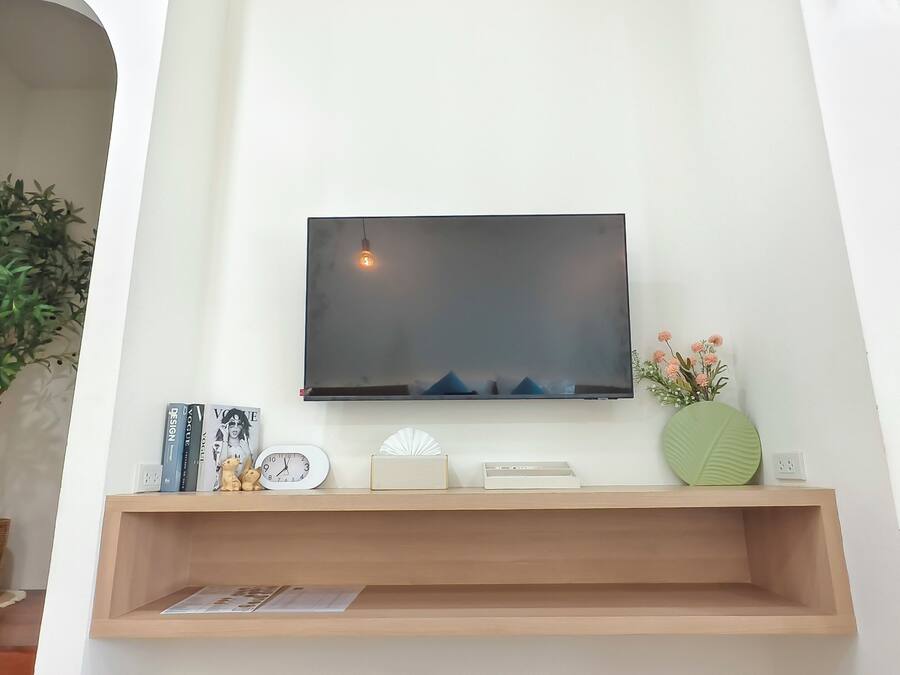 Una sala de estar con un televisor montado, un estante de madera con objetos decorativos, una planta en maceta y un reloj de pared.