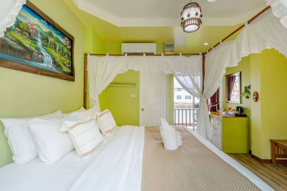 A room with a bed, a painting, an air conditioning unit, a window, and a small table with a plant.