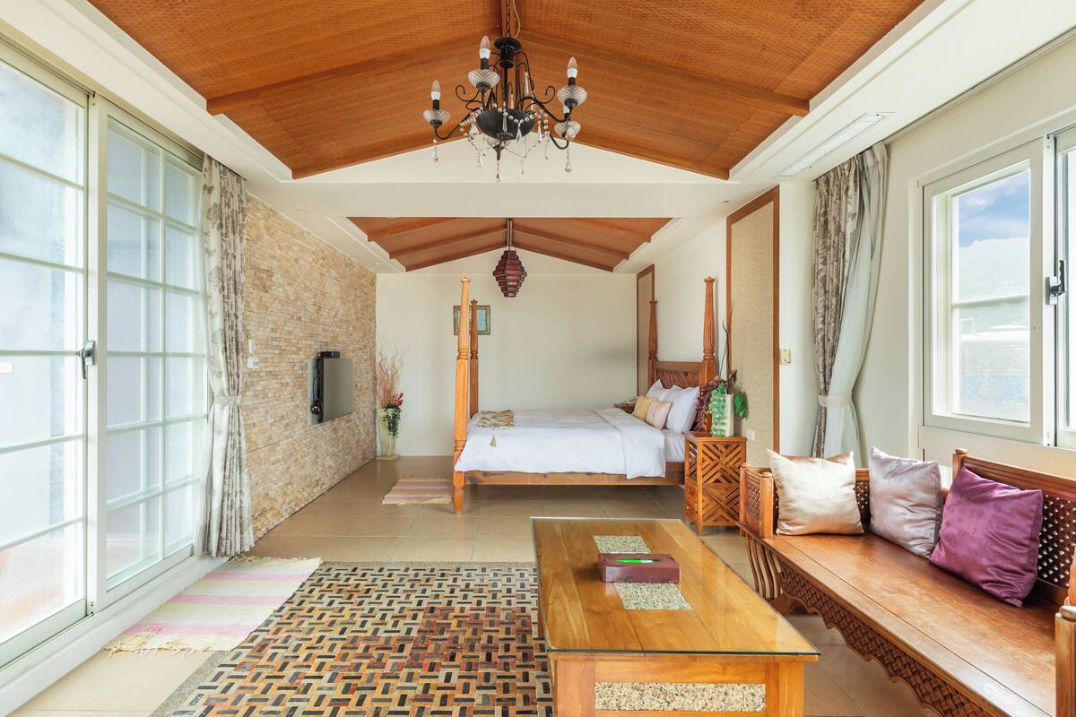 A spacious room with a large bed, a wooden bench, a glass coffee table, and a chandelier.
