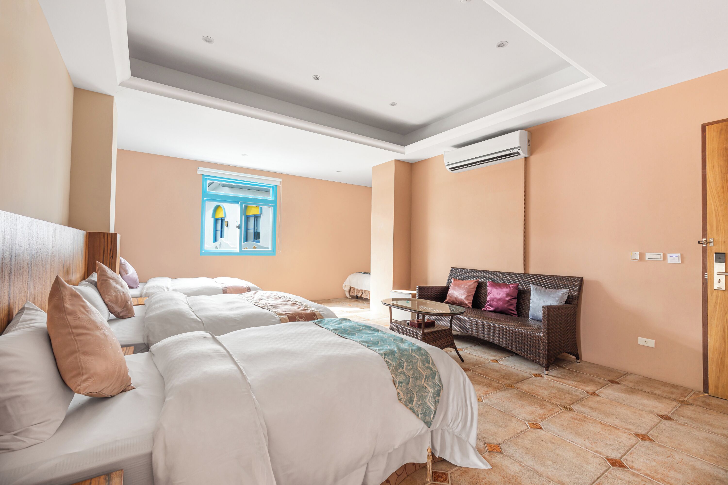 A hotel room with a large bed, a sofa, a small table, and an air conditioning unit.