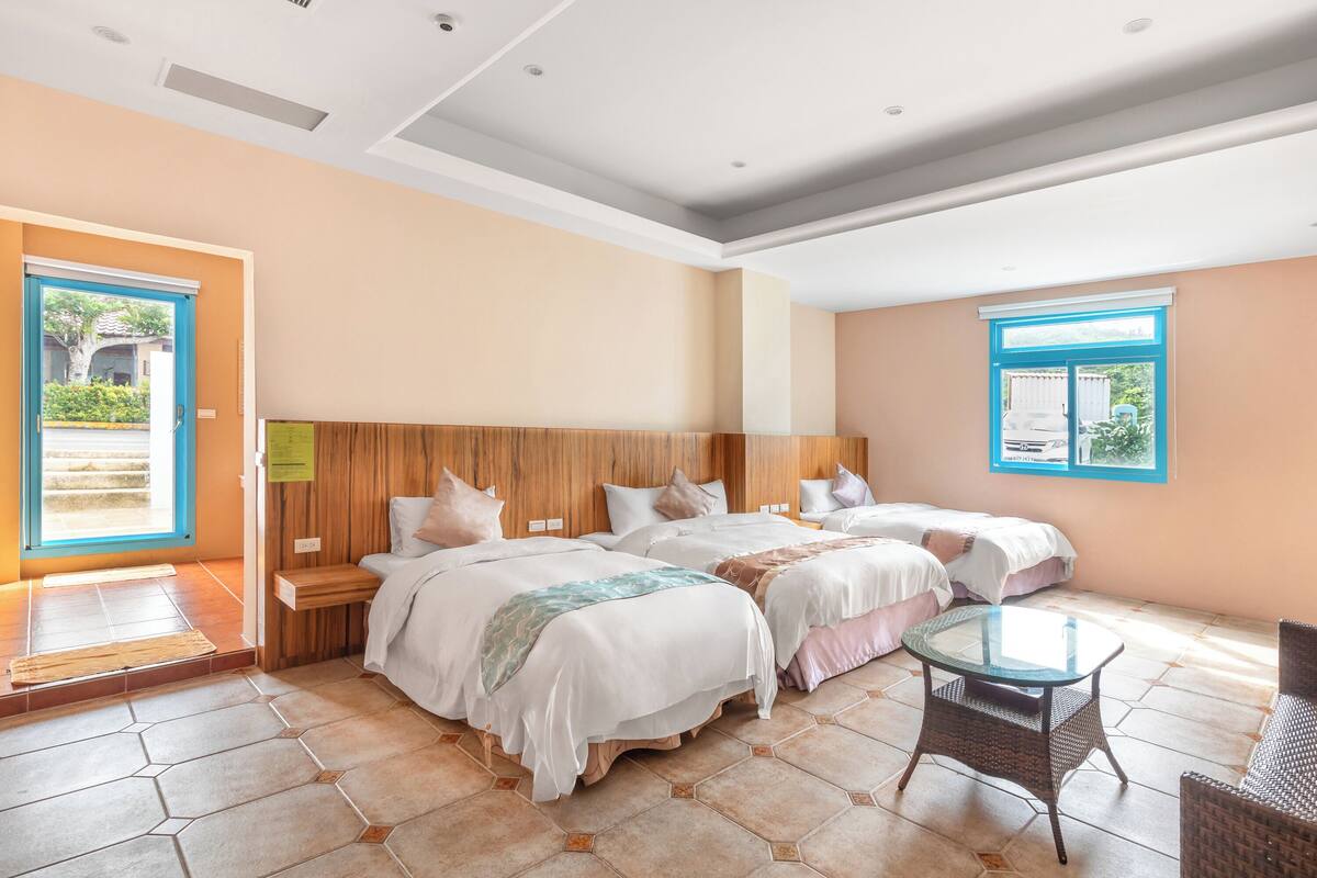 A hotel room with three beds, a glass table, and a wicker chair.