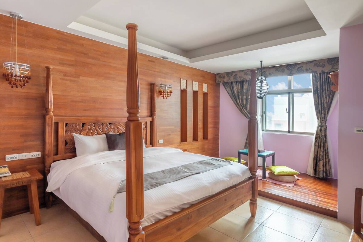 A room with a four-poster bed, wooden walls, a window with curtains, and a small table with a chair.