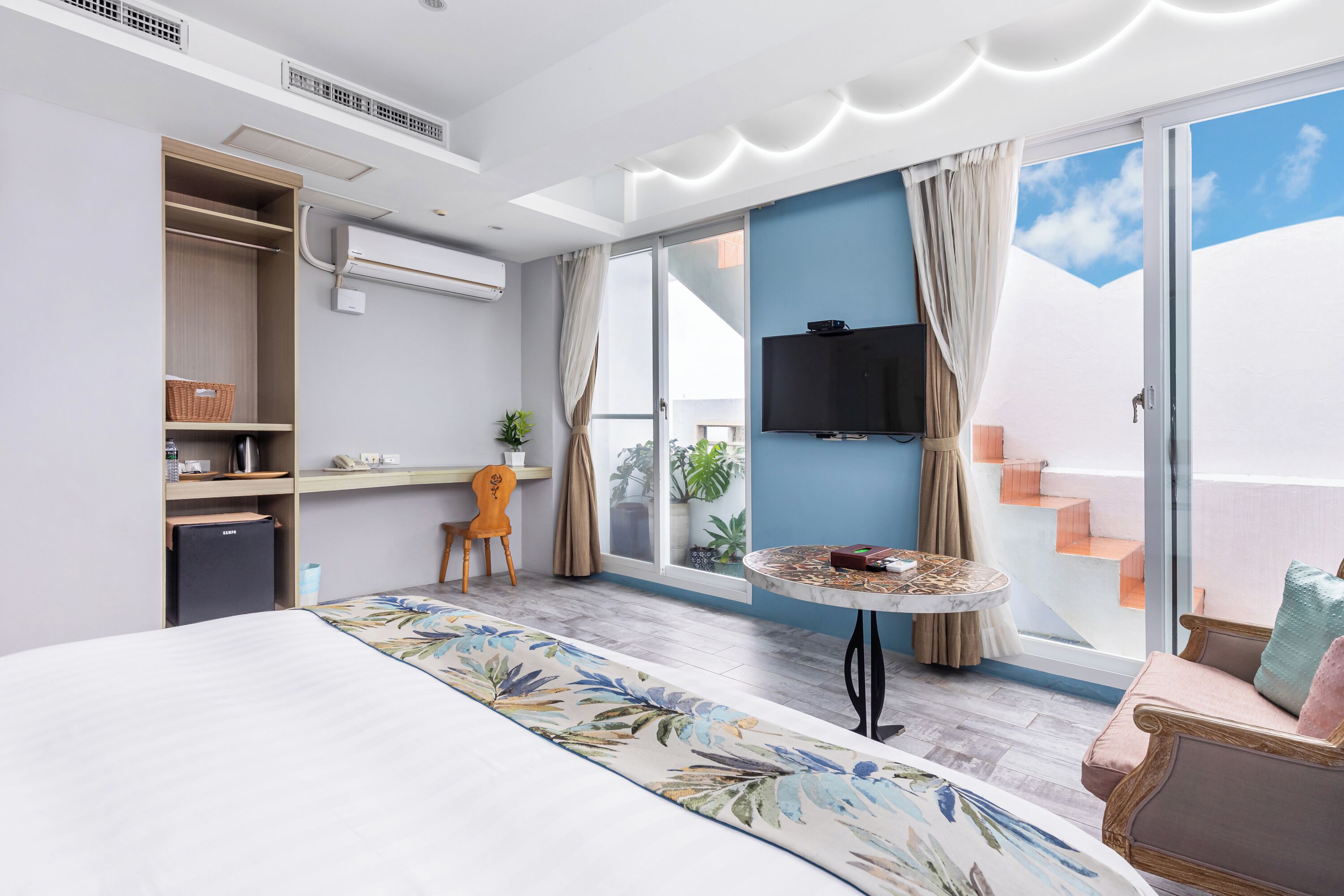 A modern hotel room with a bed, a desk, a chair, a TV, and a balcony with a view.