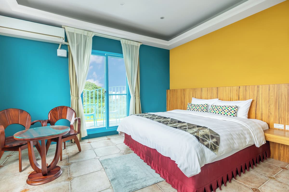 A hotel room with a bed, a wooden headboard, a dining table with chairs, and a view of the outdoors.