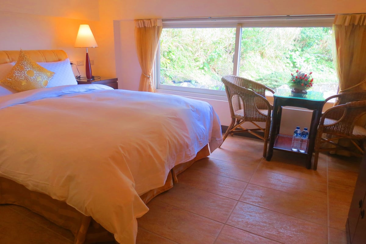 Een hotelkamer met een groot bed, een nachtkastje met een lamp, een tafel met stoelen en uitzicht op groen buiten het raam.