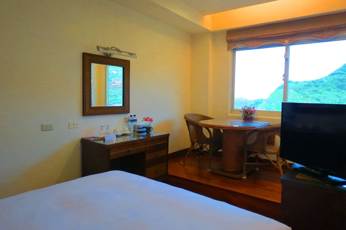 Een hotelkamer met een bed, een bureau met stoel, een televisie, een raam met uitzicht op een berg en een spiegel aan de muur.