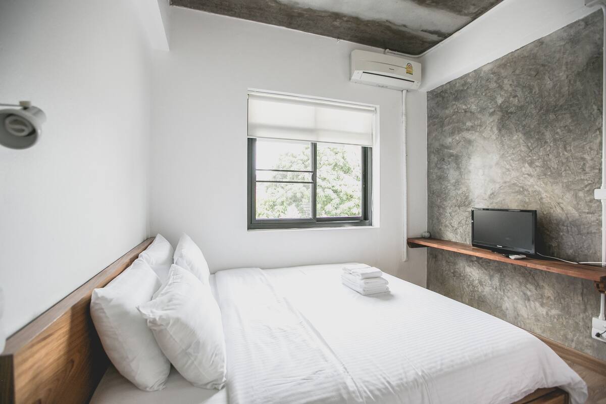 Une chambre d’hôtel avec un lit, du linge de lit blanc, une fenêtre avec des stores, un téléviseur posé sur une étagère et un appareil de climatisation.