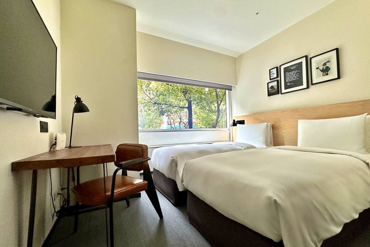 A hotel room with a bed, a desk with a chair, a window with a view of trees, and framed pictures on the wall.