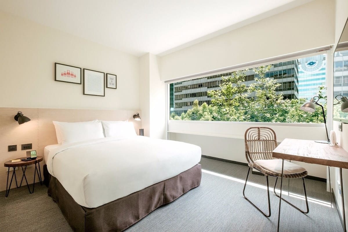 A hotel room with a bed, a desk, a chair, a window with a view of buildings and trees, and framed pictures on the wall.
