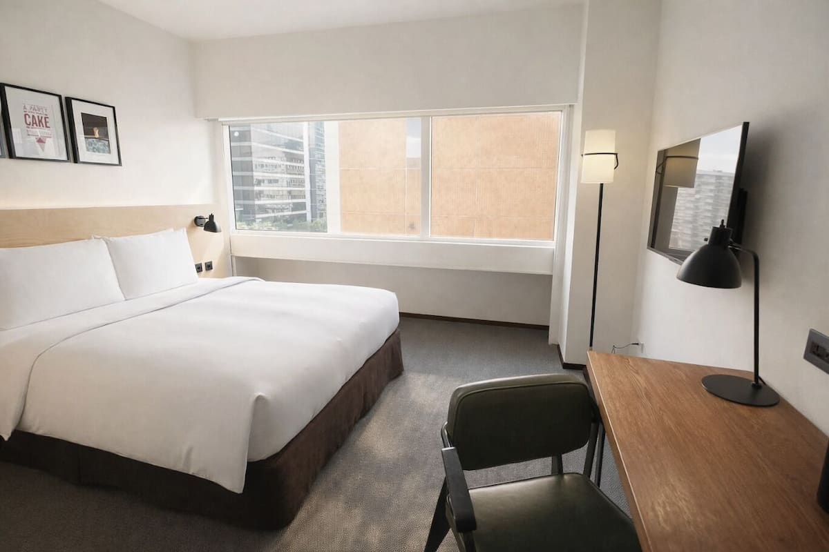 A hotel room with a large bed, a desk, a chair, a TV, a window with a city view, and framed pictures on the wall.