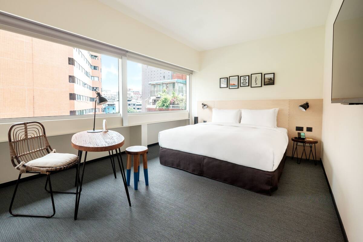 A hotel room with a large bed, a chair, a small table, and a view of the city through the window.