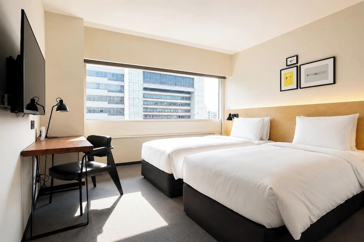 A hotel room with a desk, two beds, a TV, and a window with a city view.