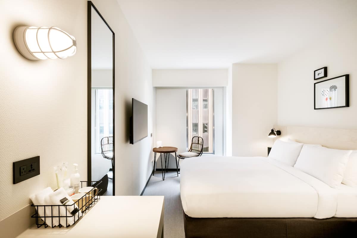 A hotel room with a large bed, a desk with a chair, a TV, and a mirror. There is a window with a view of the outside.