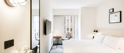 A hotel room with a large bed, a desk with a chair, a TV, and a mirror. There is a window with a view of the outside.