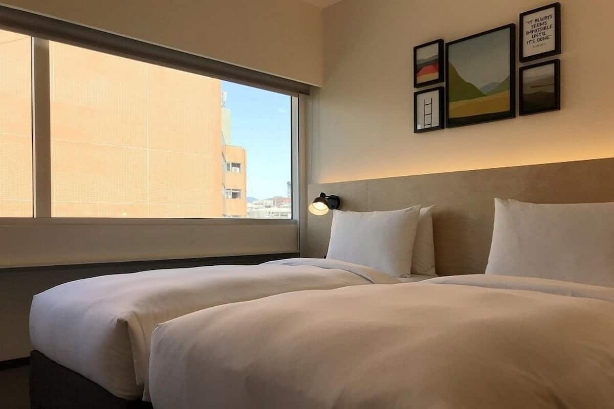 A hotel room with two beds, a large window, and framed artwork on the wall.