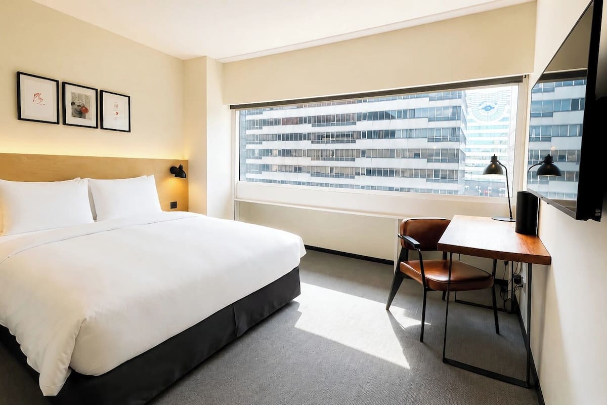 A hotel room with a bed, a desk, a chair, a TV, and a window with a city view.