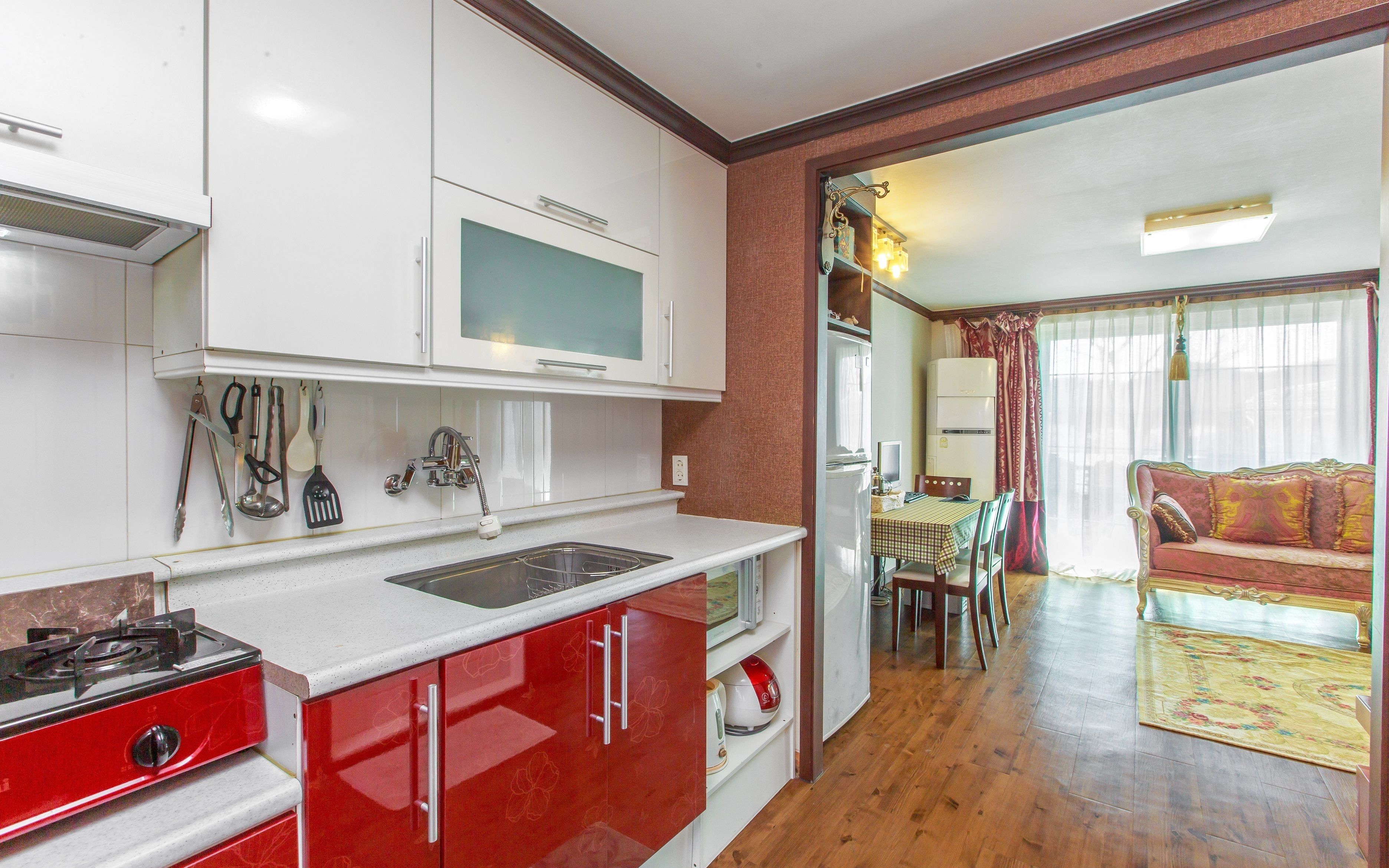 A compact kitchen with a gas stove, microwave, and sink. There is a dining area with a table and chairs, and a living room with a sofa and curtains.