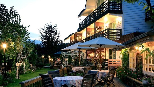 An outdoor patio with tables and chairs, surrounded by a wooden deck and lush greenery. - Spa Pension Basso (Sokcho)